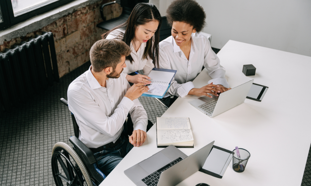staff team, one in wheelchair and two females looking at documentation with laptop on table
